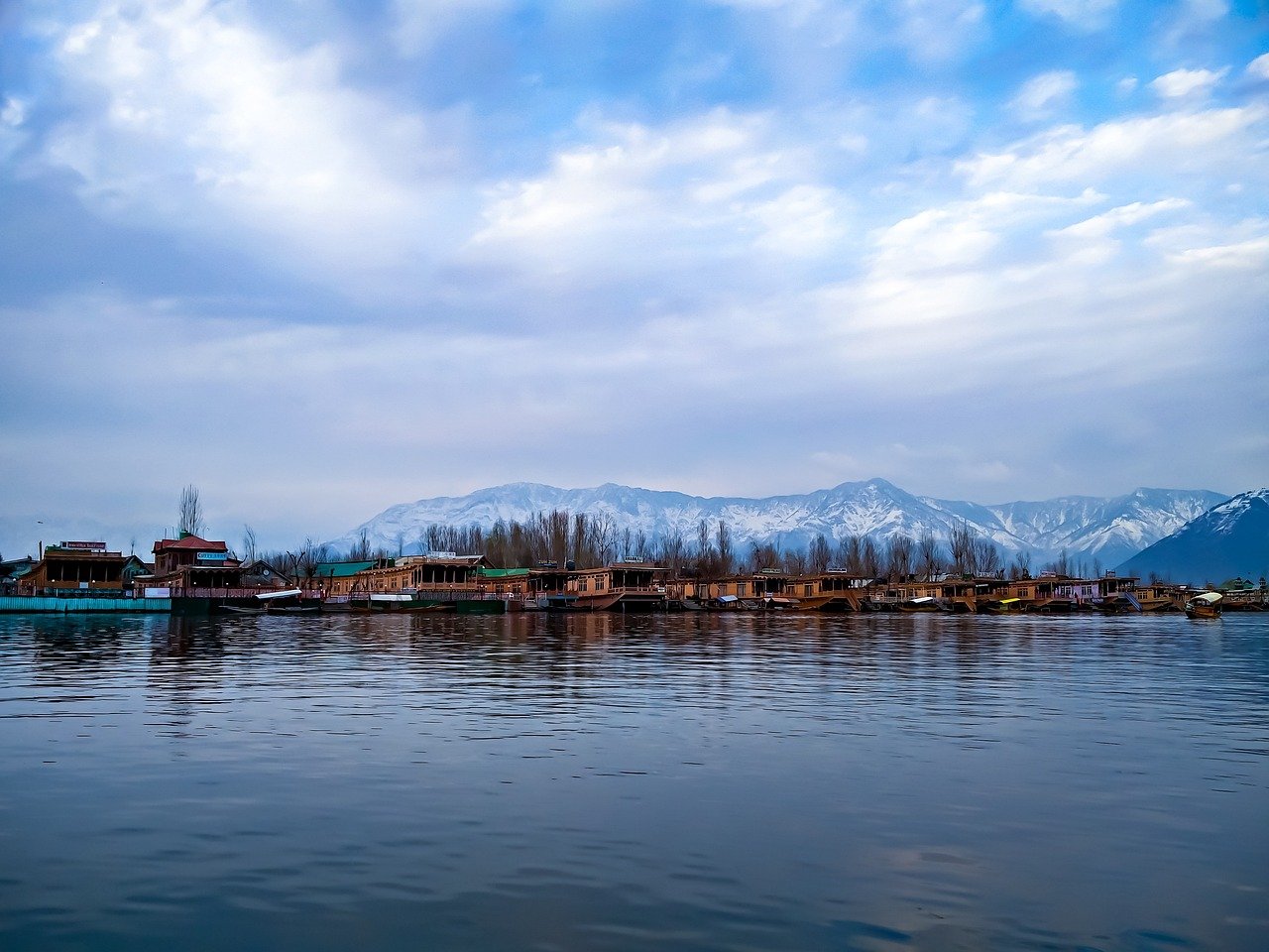 A wooden houseboat on Dal Lake Srinagar at sunrise, with carved cedar veranda, shikara tied alongside, and the Zabarwan hills behind